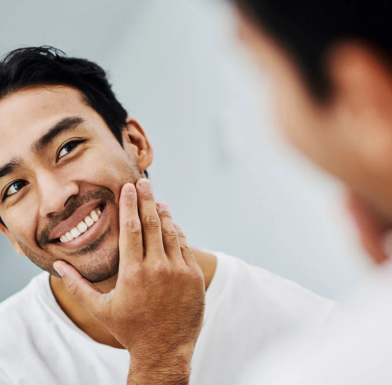 A young man with short dark hair and a beard smiles while touching his face in front of a mirror. He is wearing a white t-shirt and has a cheerful expression - Bruxism Treatment in Fort Lee, NJ