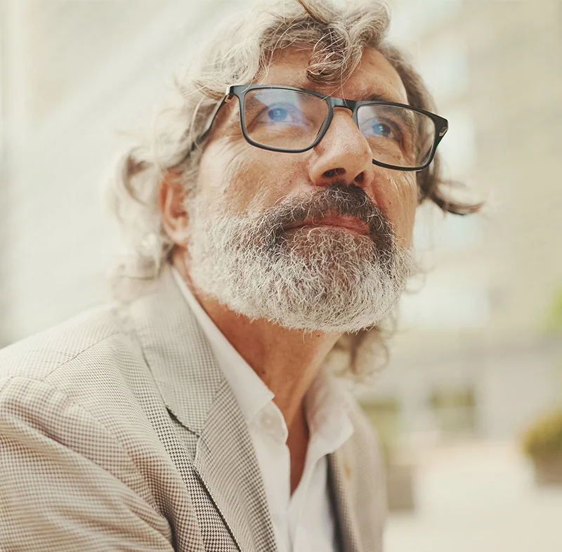A distinguished older man with gray curly hair, a beard, and glasses, looking thoughtfully into the distance while wearing a light-colored suit jacket - Oral Cancer Exams in Fort Lee, NJ