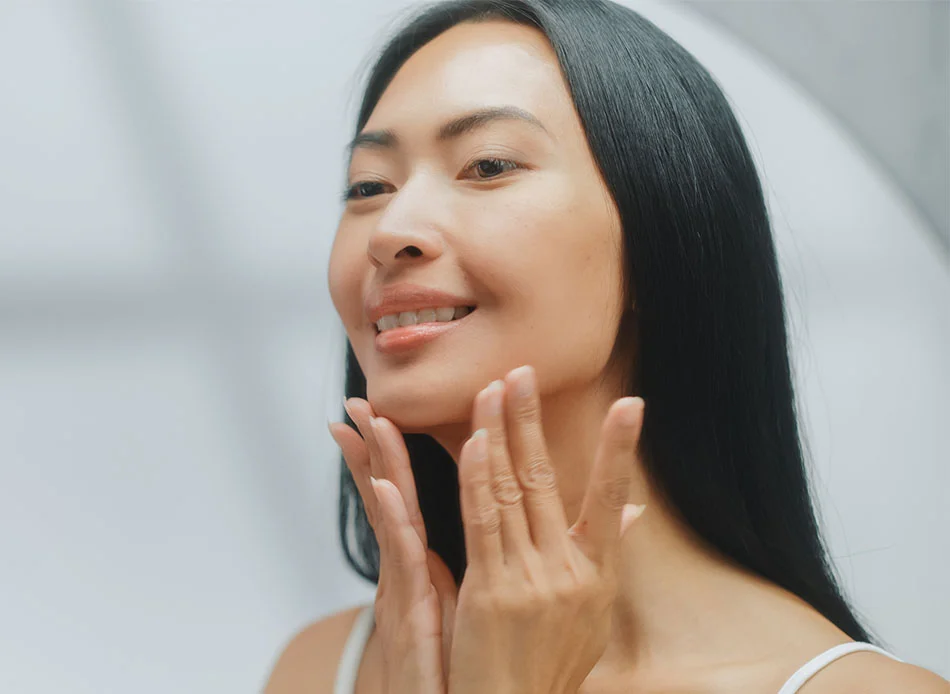 A young woman with long, straight black hair, gently touching her face with both hands, and smiling softly - Oral Cancer Exams in Fort Lee, NJ