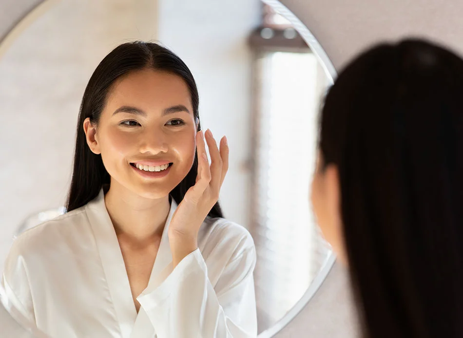woman smiling and touching her face while looking in the mirror. - Routine Dental Cleaning in Fort Lee, NJ