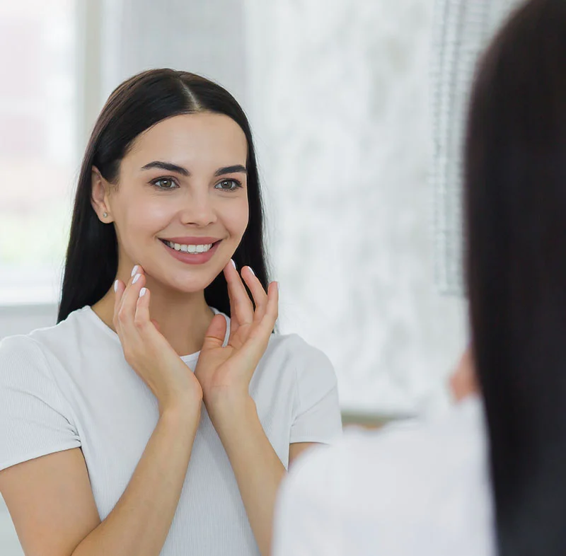 A young woman with dark hair smiles brightly as she looks at her reflection in the mirror. She wears a casual white top and gently touches her face in a well-lit, clean space - Full Mouth Debridement in Fort Lee, NJ