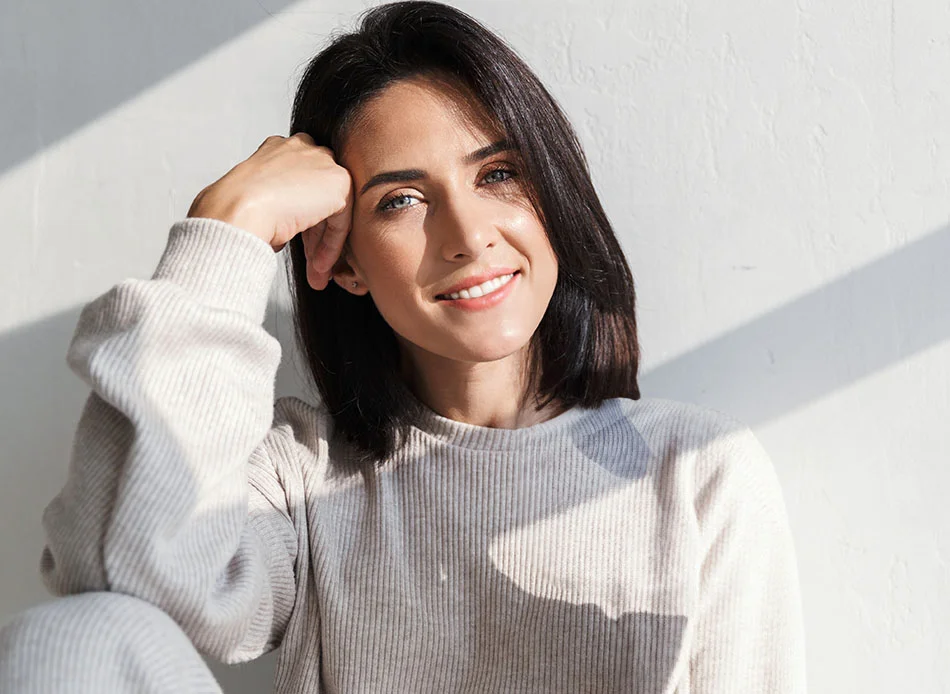A woman with short dark hair smiles confidently while resting her arm on her knee. She is dressed in a light, comfortable outfit and sits in a bright room, illuminated by natural sunlight - Full Mouth Debridement in Fort Lee, NJ