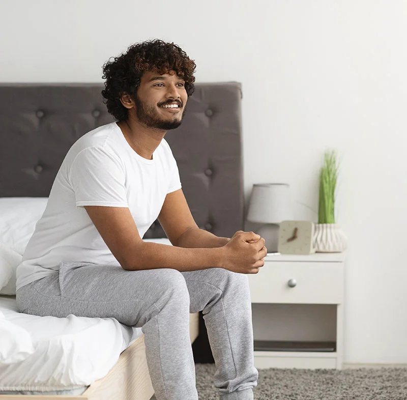 A man with curly dark hair and a beard sits comfortably on a bed in a bright, modern bedroom. He wears a white t-shirt and grey sweatpants, smiling as he looks into the distance - Initiation of Home Sleep Tests in Fort Lee, NJ