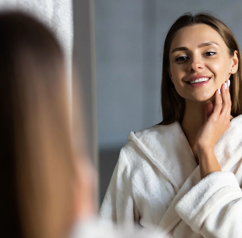 A woman with long straight hair smiles at her reflection while gently touching her face. She is wearing a plush white robe, standing in front of a mirror in a serene environment - Mandibular Advancement Devices in Fort Lee, NJ